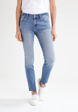 Jeans ajustados de color azul con un lavado degradado, cintura de altura media y dobladillo sin rematar en los tobillos. Combinados con zapatillas blancas sin cordones.