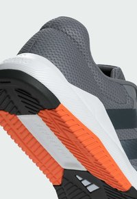 Zapatilla deportiva gris con una parte superior de malla texturizada, entresuela blanca y suela de goma con acentos naranjas y un patrón de pisada distintivo.