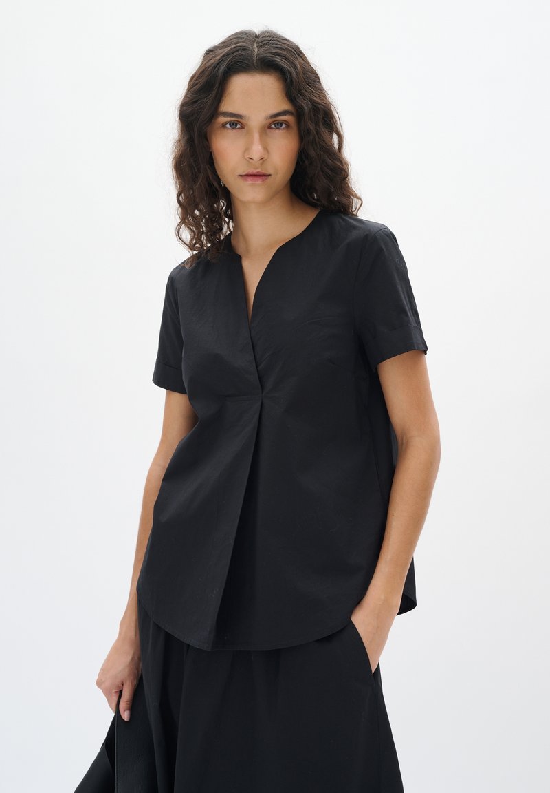 Top nero a maniche corte con scollatura a V e dettagli arricciati nella parte anteriore. Tessuto leggero con una texture liscia, silhouette aderente.