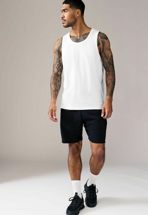 Weißes Tanktop, schwarze Shorts, schwarze Sneakers und weiße Socken. Modell mit Tattoos steht vor einem neutralen Hintergrund, der das Outfit hervorhebt.