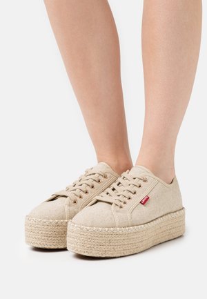 Beige Leinenschuhe mit Plattformsohle aus geflochtenem Jute, runden Schnürsenkeln, kupferfarbenen Ösen und einem kleinen roten Logoetikett an der Seite.