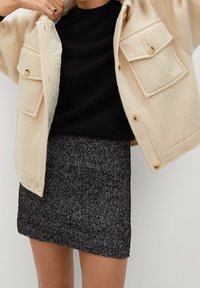 Femme portant un manteau beige à boutons sur un haut noir et une jupe à motif chevrons gris foncé.