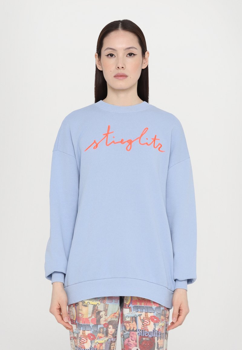 Light blue oversized sweatshirt with a crew neckline and coral embroidered text "stiglitz" across the chest. Soft cotton fabric with a relaxed fit.