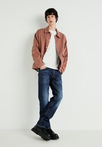 Chaqueta ligera de color óxido sobre una camiseta blanca, combinada con jeans de mezclilla azul oscuro y zapatos negros con cordones de suela gruesa. Diseño minimalista.
