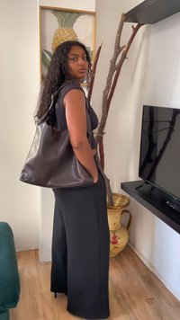 Mulher com longos cabelos cacheados, vestindo uma blusa preta sem mangas e calças, carregando uma grande bolsa de couro marrom-escuro, numa sala com uma televisão e um vaso decorativo.