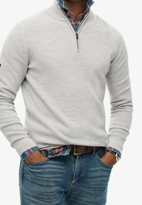 Pull-over gris clair avec une fermeture éclair au quart, manches longues, texture côtelée, porté par-dessus une chemise à carreaux aux nuances de bleu, vert et rouge.