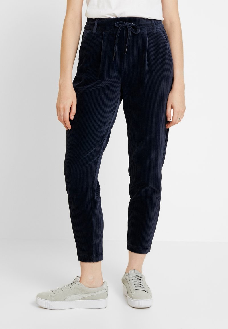 Femme portant un pantalon en velours côtelé noir avec une taille à cordon et des baskets blanches sur un fond uni.