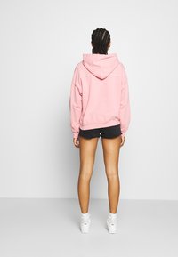 Vaaleanpunainen huppari, jossa on rento istuvuus, kenguru-tasku ja kiristysnyörillinen huppu, yhdistetty mustiin farkkushortseihin ja valkoisiin lenkkareihin.