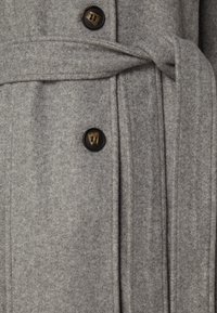 Cappotto di lana grigio con due bottoni marrone scuro e una cintura legata in vita, che mostra un tessuto testurizzato e cuciture a vista.