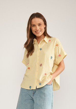 Naeratav naine pikkade pruunide juustega, kandes kollase ruudulise lühikeste varrukatega särgi tikitud kalade ja koostratega, sobitatud helesiniste teksadega.