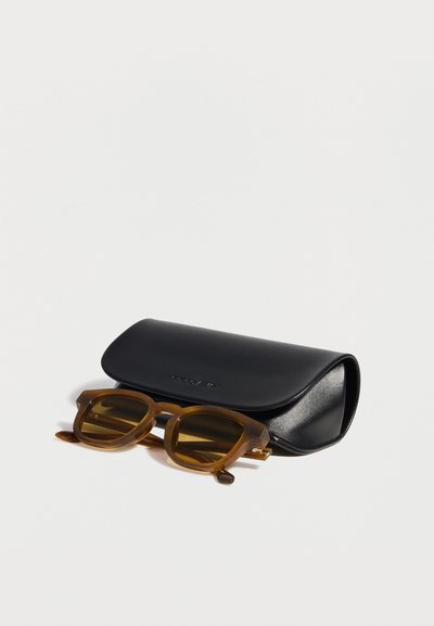 Lunettes de soleil carrées translucides marron avec des verres sombres posées devant un étui à lunettes en cuir noir fermé sur une surface blanche.