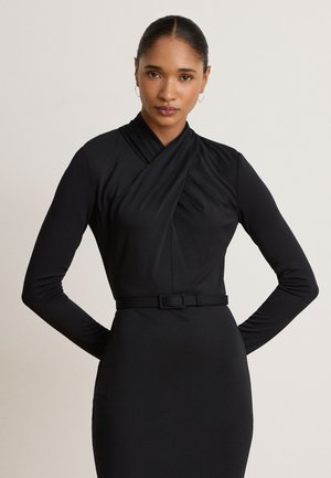 Femme aux cheveux foncés en chignon lisse, portant une robe noire cintrée à manches longues avec une taille ceinturée et un décolleté croisé, debout, les mains dans le dos.
