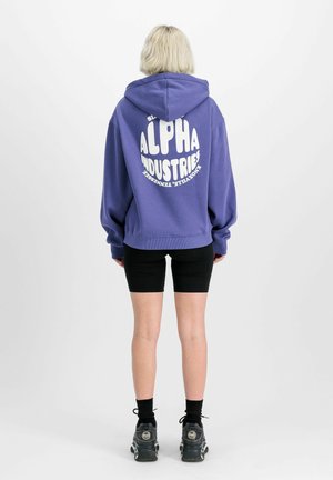 Purple hoodie with white graphic text on the back, fitted black shorts, and black sneakers, featuring a round logo and relaxed fit.