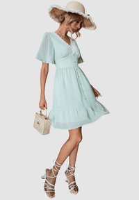 Un vestido verde menta con mangas cortas y vaporosas, cuello de encaje, botones en el frente y dobladillo con volantes, combinado con sandalias blancas de tiras y un bolso de mimbre.