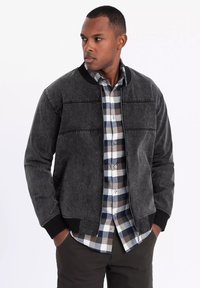 Harmaa pompputakki, jossa on ribbikaulus ja hihansuut, sileä pinta ja kaksi vaakasuoraa rintaommelta, päällä ruudullinen paita.