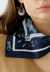 Navyblauwe sjaal met witte paisley- en bloempatronen, afgezet met witte stiksels, gecombineerd met een zilveren halfmaansvormige oorbel.