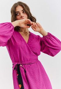 Różowa sukienka kopertowa z bufiastymi rękawami, wzór w grochy oraz czarny pasek z kolorowymi akcentami. Gładka tkanina, dekolt w serek.