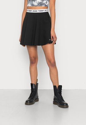 Kvinde iført en sort udvidet nederdel med en hvid logo-talje, sorte snørelæder-støvler og en cropped tie-dye top, stående op ad en hvid væg.