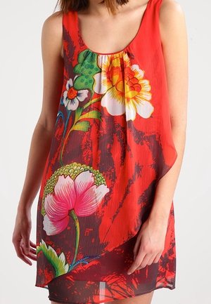 Femme portant une robe rouge sans manches avec de grands motifs floraux colorés en jaune, rose, blanc et vert sur le devant, sur fond blanc.