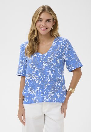 Ženska z dolgimi blond lasmi nosi modro-belo cvetlično bluzo z V-izrezom in bele hlače, nasmejana, z eno roko v žepu.