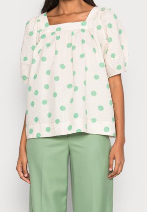 Femme portant un chemisier crème à manches courtes avec des pois verts et un col carré, associé à un pantalon taille haute vert clair.