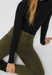 Jean ajusté vert olive en denim, doté d'une braguette zippée et d'un design à cinq poches, associé à un haut noir à manches longues.
