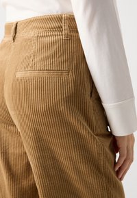 Close-up van een persoon die tan corduroy broeken draagt met een achterzak en een witte lange mouwen shirt manchet zichtbaar nabij de hand.