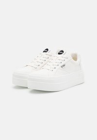 Bílé nízké platformové tenisky s tkaničkami, kovovými očky, strukturálními silnými podrážkami a černými logovými nášivkami na jazyku a na straně.