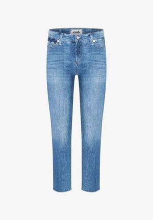 Femme portant un jean skinny court bleu clair avec des ourlets effilochés et des ballerines à enfiler à motifs, vue de dos sur un fond uni.