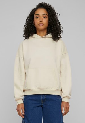Beige hettegenser laget av myk stoff. Den har en kengurulomme, hette med snøring og elastiske mansjetter. Kombinert med blå denim bukser.