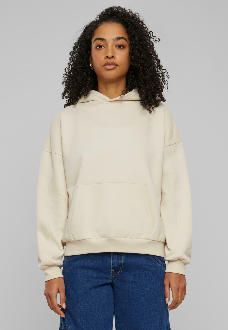 Beige hettegenser laget av myk stoff. Den har en kengurulomme, hette med snøring og elastiske mansjetter. Kombinert med blå denim bukser.