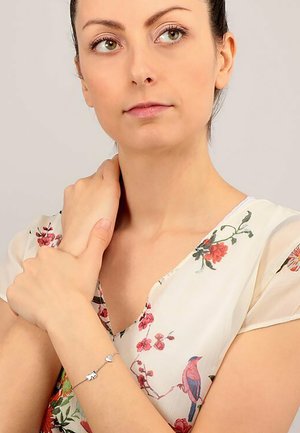 Silberarmband mit Tier- und Herzanhängern, getragen zu einer blusenartigen Oberteil mit leichtem Floralprint aus zarter, transparenter Stoff in Pastellfarben.