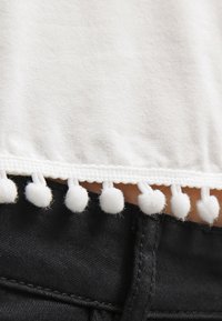 Top in tessuto bianco con orlo testurizzato adornato da piccoli pompon bianchi, abbinato a un materiale in denim nero visibile sotto.