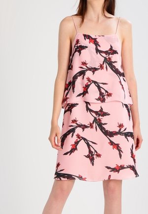 Vrouw die een roze mouwloze jurk draagt met rood-zwart bloemenpatroon, staand tegen een effen lichte achtergrond.