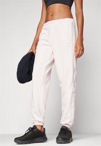Lichtroze joggingsbroek met een elastische tailleband, zijstrepen en logo-detail; gecombineerd met zwarte mesh sneakers en een donker accessoire.