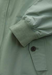 Veste verte avec une finition mate et lisse. Comprend des poignets élastiques, une poche à bouton, et un design épuré avec des détails de couture minimalistes.