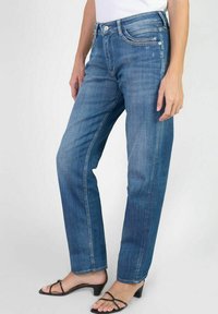 Jeans en denim bleu moyen avec une coupe droite, cinq poches et un léger effet de décoloration. Portés avec des sandales à talons noires.