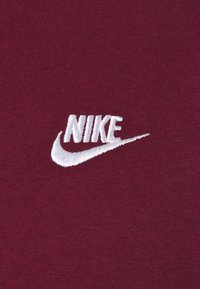 Vitt broderat Nike-logotyp med swoosh på mörkröd tygbakgrund.