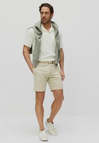 Avslappnad outfit bestående av en ljusgrå tröja draperad över en vit kortärmad polo och beigea shorts, tillsammans med vita sneakers.