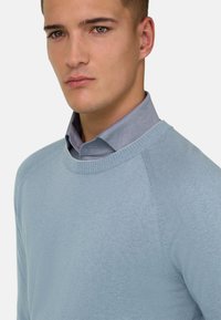 Jeune homme aux cheveux courts portant un pull bleu clair à col rond par-dessus une chemise bleue à col, expression neutre, sur un fond uni.