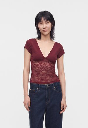 Jeune femme aux cheveux noirs portant un haut en dentelle bordeaux à manches courtes et un jean bleu foncé, debout devant un fond uni.