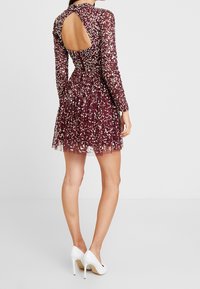 Kvinna som bär en kort bourgognyklänning med paljetter och en tårformad ryggöppning, ihop med vita högklackade skor, stående mot en enfärgad bakgrund.