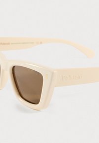 Ανοιχτό μπεζ ρετρό γυαλιά ηλίου με τριγωνικούς σκελετούς, καφέ φακούς, ανάγλυφο λογότυπο "Polaroid" στους βραχίονες και τρία μικρά χρυσά διακοσμητικά στην μπροστά πλευρά.