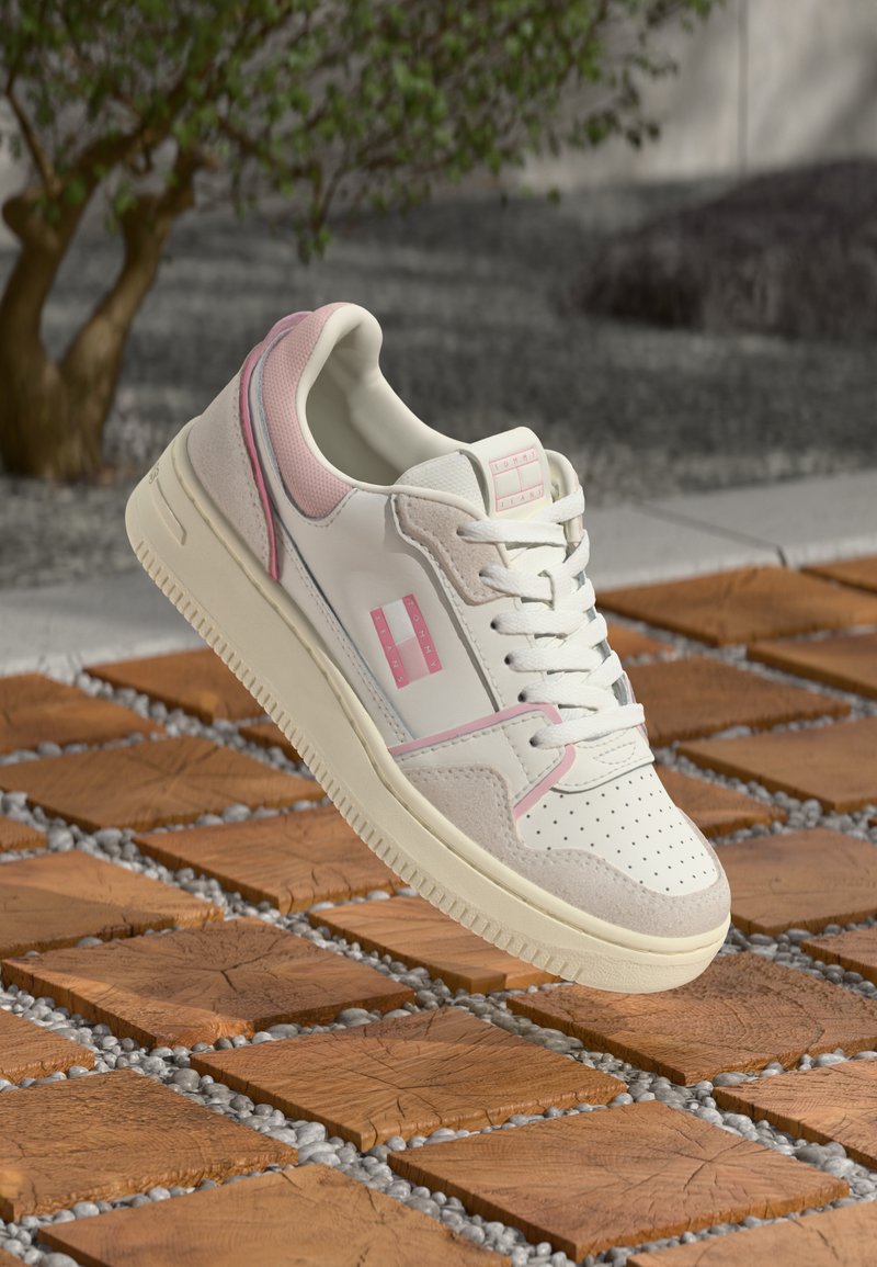 Witte en lichtroze Tommy Jeans sneaker met veters, geperforeerde neus en suède accenten, zwevend boven houten tegelvloer in de buitenlucht.