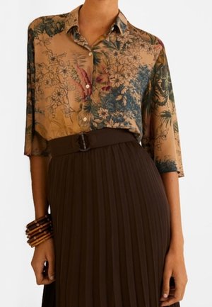 Przejrzysta kwiecista bluzka w beżowym i zielonym kolorze z kołnierzykiem, guzikami z przodu i rękawami do połowy przedramienia, połączona z plisowaną brązową spódnicą.