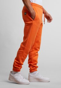 Orange Sweatpants mit Taschen, elastischem Bund und Bündchen. Kombiniert mit weißen Sneakers, die ein sichtbares Logo auf den Hosen haben.