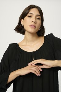 Schwarze Bluse mit Rundhalsausschnitt und Knopfdetails, mit plissiertem Design und langen Ärmeln. Model trägt ein silbernes Armband am Handgelenk.