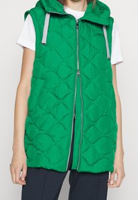 Gilet sans manches matelassé vert avec un col haut, fermeture éclair à l'avant et sangles d'accent gris. Texture lisse avec un design léger et rembourré.