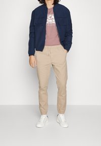 Veste bleue marine avec deux poches poitrine, t-shirt rose clair avec texte, pantalon beige fuselé et baskets blanches avec accents noirs.