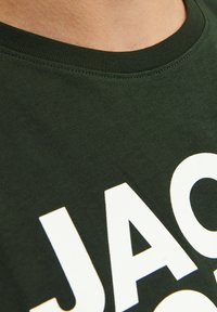 Camiseta de algodón verde oscuro con cuello redondo y grandes letras blancas. La tela parece suave y tiene una textura blanda.